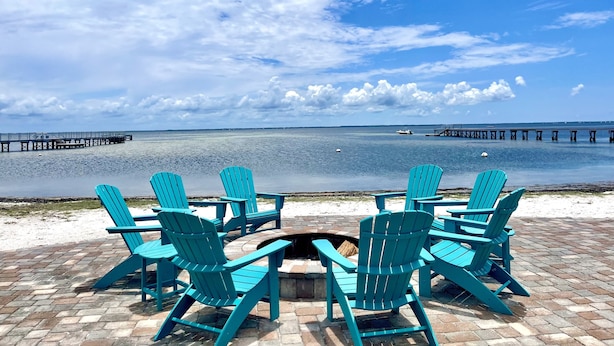 Uma área de estar à beira-mar com cadeiras Adirondack turquesa voltada para um mar calmo e um píer ao longe.