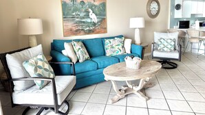 A living room with a blue sofa, two armchairs, a coffee table, and a painting of a bird on the wall. - Surf Side Shores 2405  2 bd 2 ba  gulf-front  (Gulf Shores)