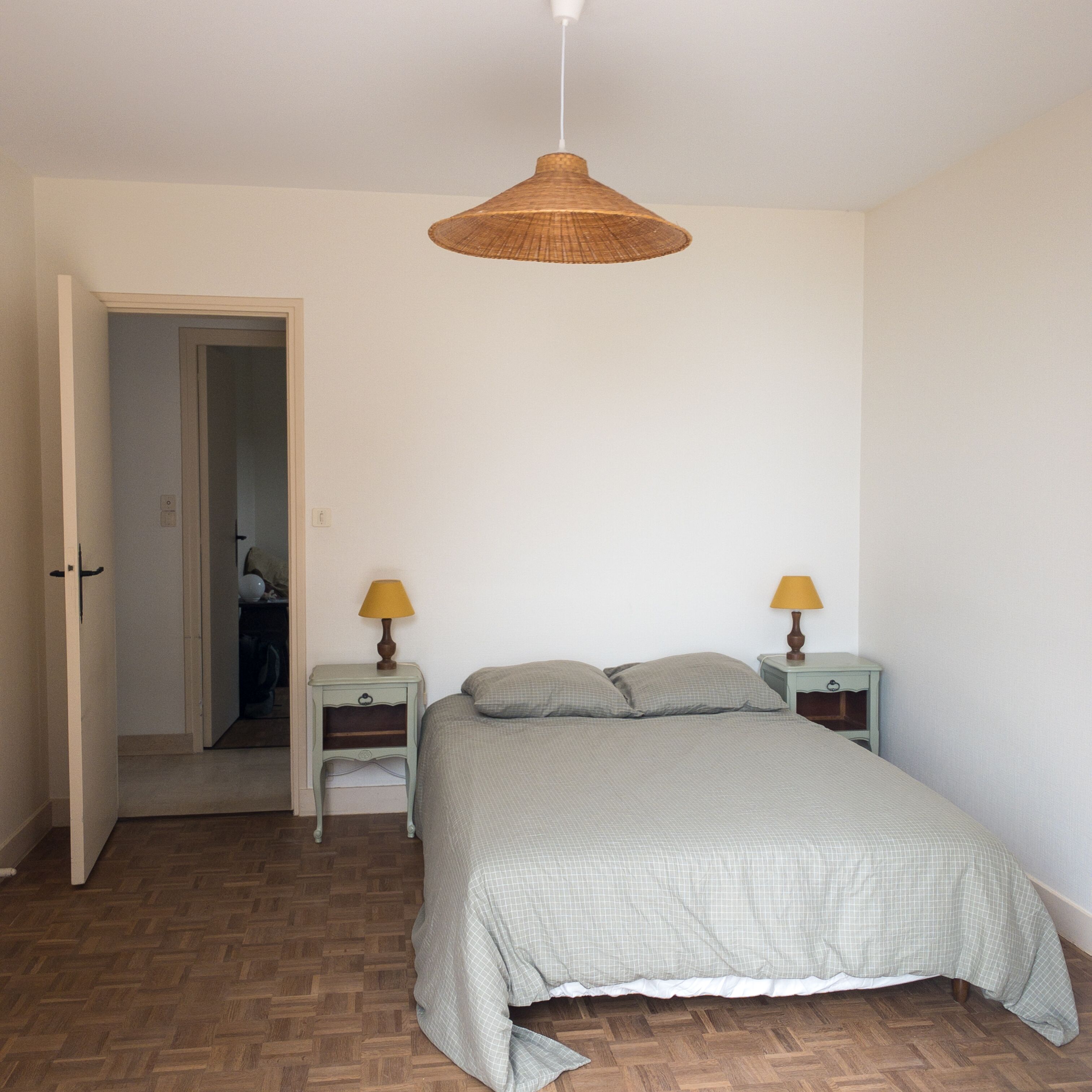 Een slaapkamer met een bed, twee nachtkastjes met lampen, een deur naar een andere kamer en een houten vloer met een visgraatmotief.