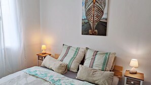 Een opgemaakt bed met streepjesbeddengoed, twee nachtkastjes met lampen, en een aan de muur hangend schilderij van een boot.