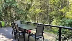 Une terrasse en bois, agrémentée d’une table et de chaises, entourée d’une rambarde en métal, et offrant une vue sur une zone boisée.