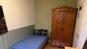 A small bedroom with a single bed, a wooden wardrobe, and a green cushioned stool.