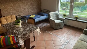 A small, cozy room with a bed, a chair, a table with a plant, and a view of a green outdoor area through a window.