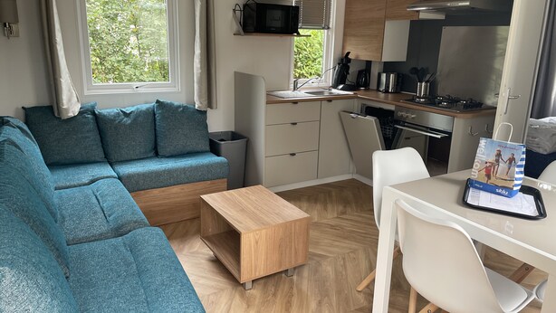 Een ruimtelijk ingerichte leefruimte met een bank, een eettafel met stoelen, een keukenhoek met een magnetron en uitzicht op groen.