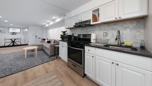 Una cocina moderna con armarios blancos, una estufa de acero inoxidable y un horno empotrado. Hay una zona de comedor con una mesa y sillas, y una alfombra gris en el suelo.
