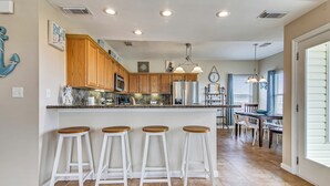 A modern kitchen with wooden cabinets, a central island with stools, and a dining area with a table and chairs. - Sunflower Surf by Ryson Vacation Rentals (Galveston)
