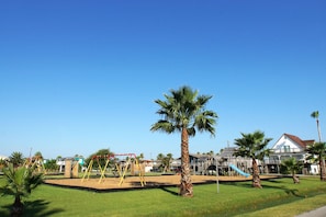 A playground with swings and a slide, surrounded by palm trees and a building with a balcony. - Sunflower Surf by Ryson Vacation Rentals (Galveston)