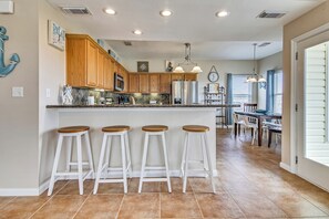 A modern kitchen with wooden cabinets, a central island with stools, and a dining area with a table and chairs. - Sunflower Surf by Ryson Vacation Rentals (Galveston)
