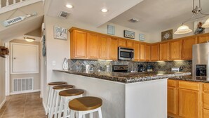 A kitchen with wooden cabinets, a granite countertop, and a stainless steel refrigerator. There are three white bar stools and a microwave. - Sunflower Surf by Ryson Vacation Rentals (Galveston)