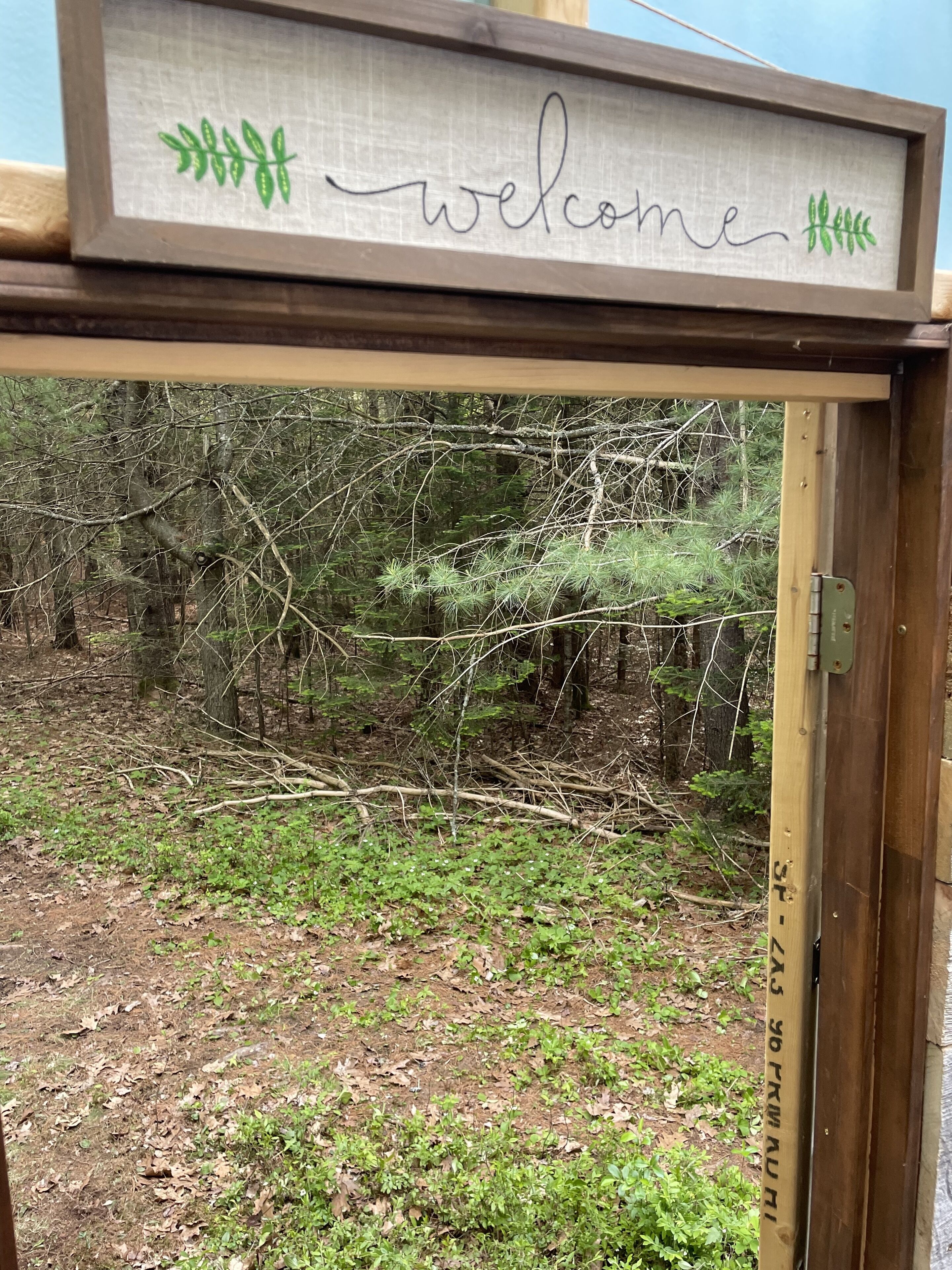 En träskylt med ordet ”välkommen” och gröna lövmotiv, placerad mot en bakgrund av en skog.