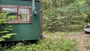 Uma pequena cabana de madeira verde, com janela e chaminé de metal, rodeada por uma floresta densa.