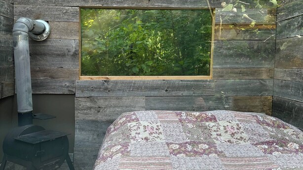 Quarto de madeira rústico, com uma cama, uma janela pequena com vista para o verde e um fogão a lenha.