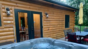 Una cabaña de madera con un jacuzzi en primer plano, un comedor con sillas y una mesa, y un paraguas amarillo.