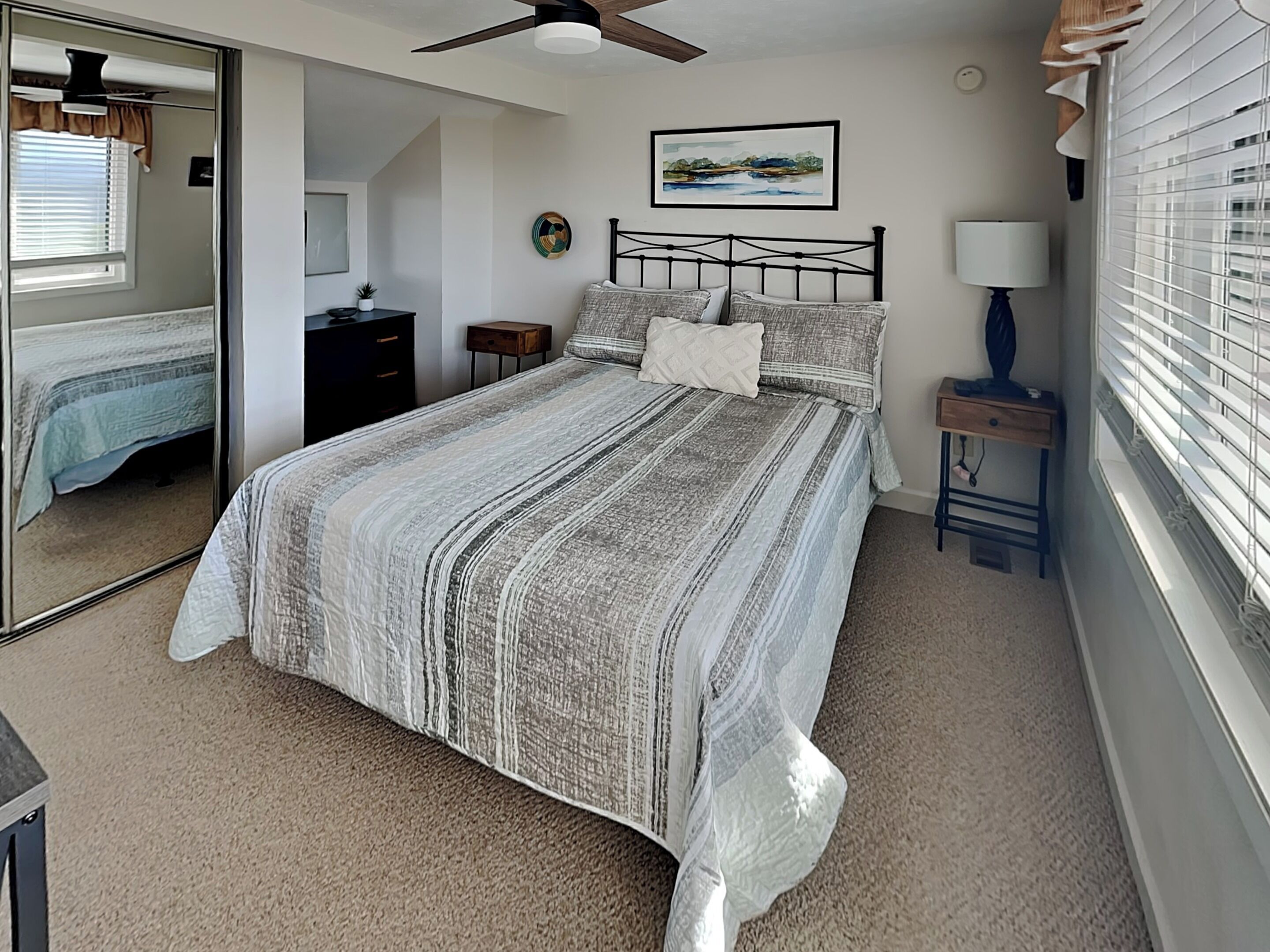 A bedroom with a bed, a nightstand, a lamp, a mirror, a window with blinds, and a ceiling fan.
