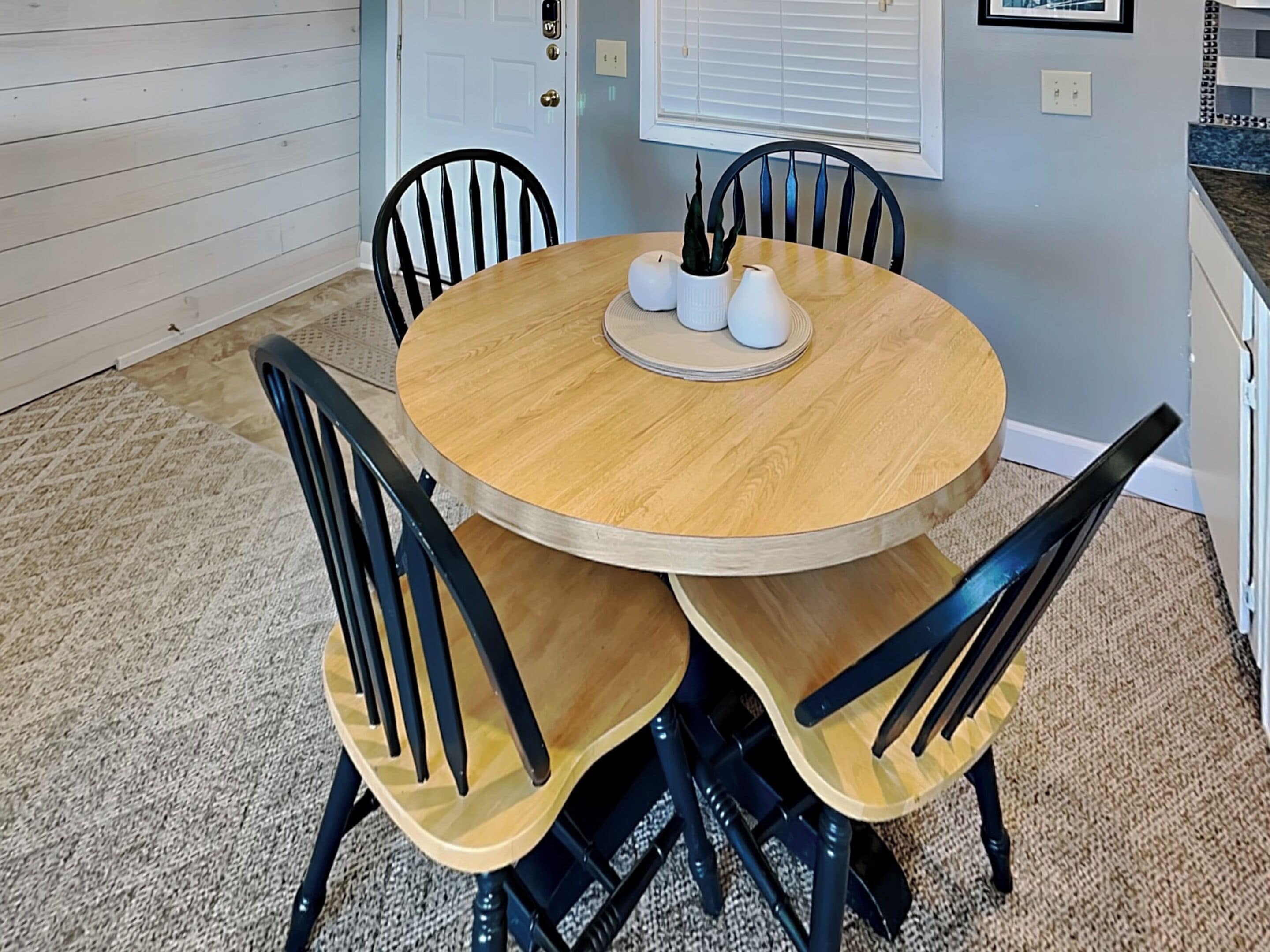 A round wooden dining table with four black chairs, a white vase, and a small potted plant on the table.