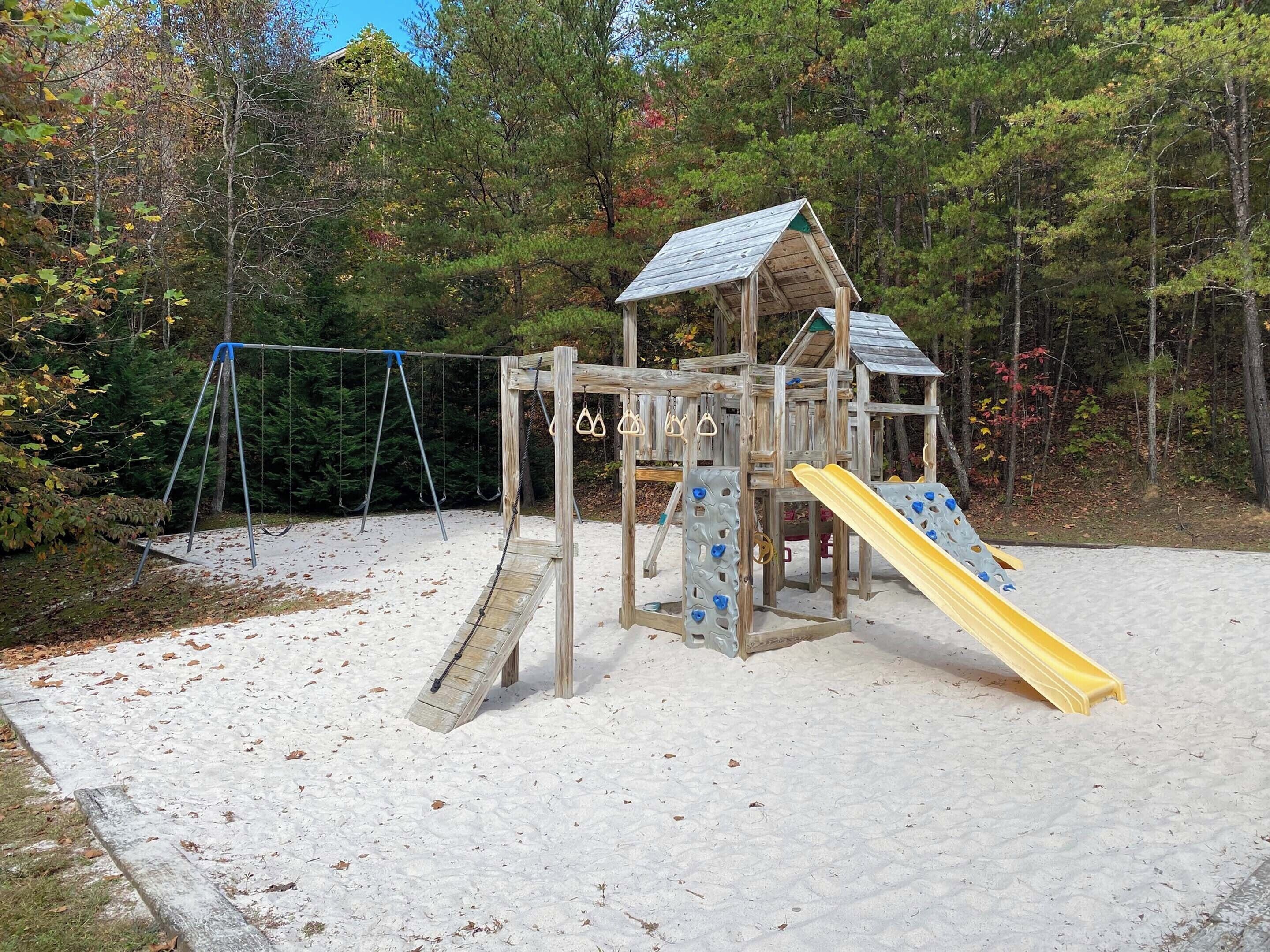 A wooden playground with a slide and climbing wall, surrounded by trees.