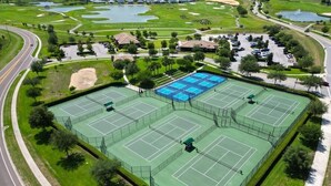 A large tennis court complex with multiple courts, surrounded by greenery and a parking area.