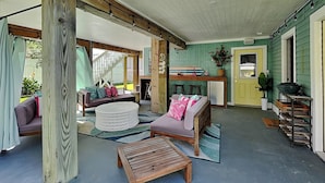 A covered outdoor living area with a wooden bench, a round white ottoman, and a sofa with cushions. - The Blue Flamingo by Ryson Vacation Rentals (Galveston)