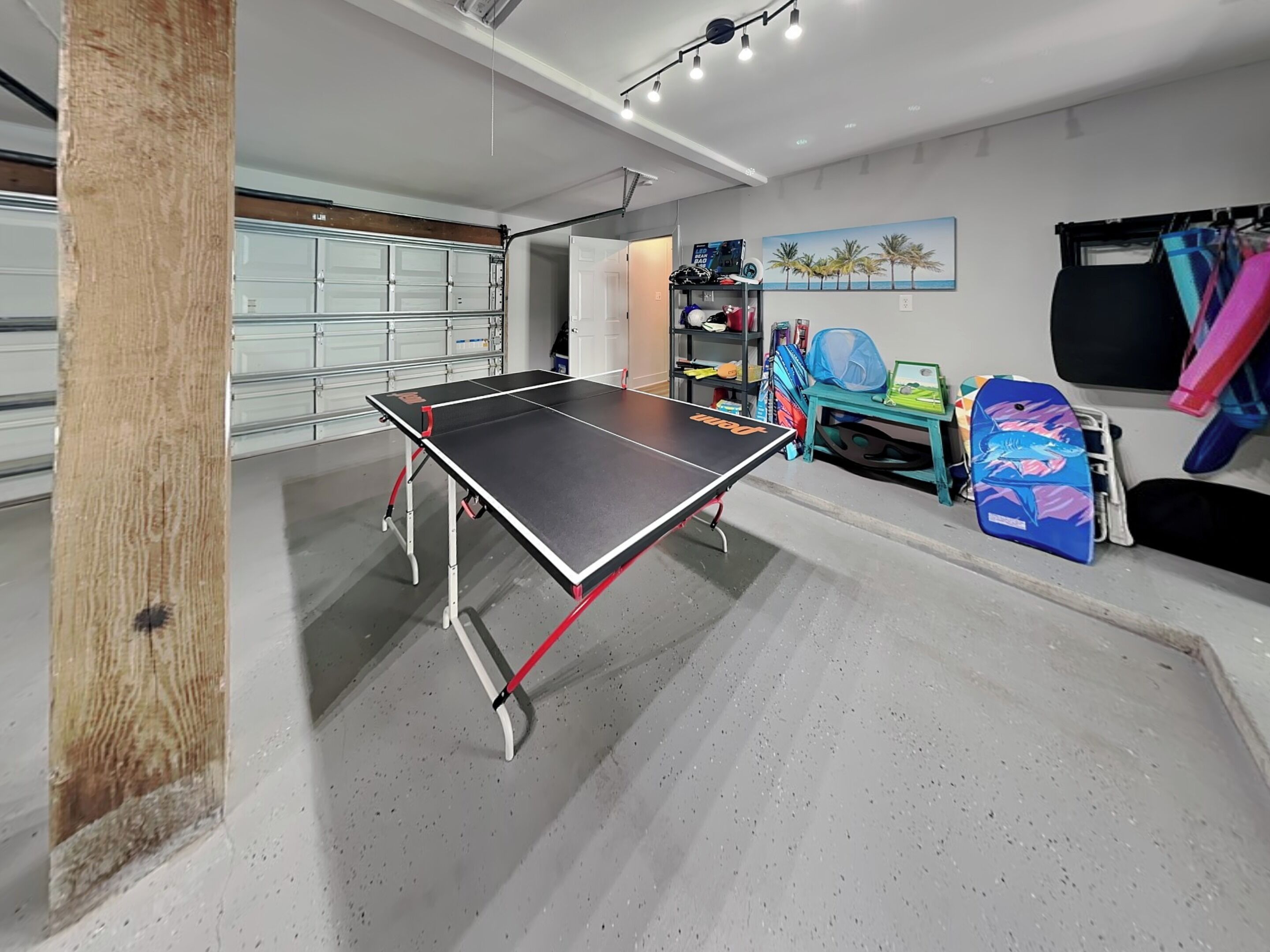 A ping pong table in a room with a garage door, shelves with items, and a painting on the wall.
