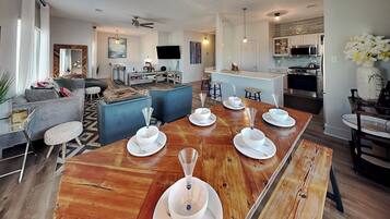 Una cocina moderna con una isla central, un comedor con una mesa de madera preparada para seis personas y una sala de estar con un sofá y un televisor.