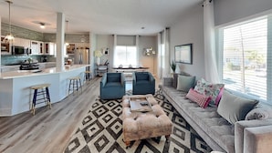 A modern living room with a sofa, ottoman, and coffee table, a kitchen area with a bar, and a dining space. - The Blue Flamingo by Ryson Vacation Rentals (Galveston)