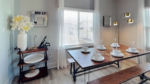A dining area with a wooden table set for four, a vase with flowers, a mirror, and a floor lamp. - The Blue Flamingo by Ryson Vacation Rentals (Galveston)