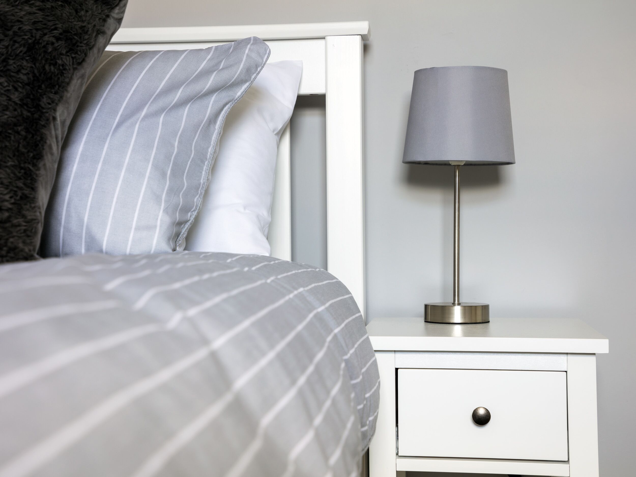 A neatly made bed with striped bedding, a white bedside table with a lamp, and a drawer.