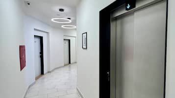 Un pasillo moderno con suelo de baldosas, un letrero de salida de emergencia rojo y un ascensor mural con una flecha direccional.