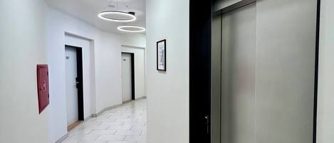 A modern hallway with a tiled floor, a red emergency exit sign, and a wall-mounted elevator with a directional arrow.