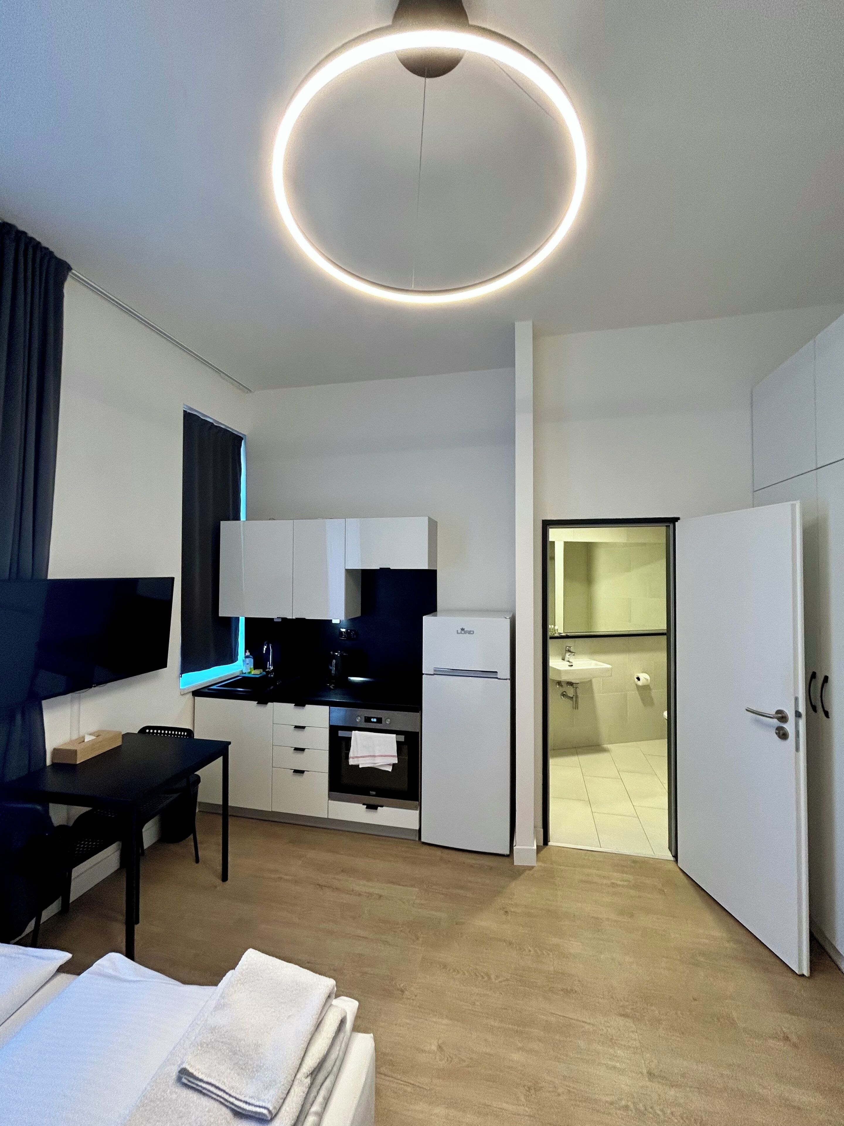 A modern, well-lit room with a round ceiling light, a flat-screen TV, a kitchenette, a dining area, and a bathroom.