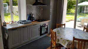 Een keuken met een spoelbak, fornuis en kasten, een eettafel met stoelen, en uitzicht op een tuin door glazen deuren.