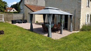 Een overdekt terras met een tafel en stoelen, een hond die op het gras ligt, en een stenen gebouw op de achtergrond.