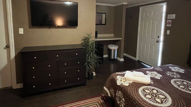 A hotel room with a dark wooden dresser, a flat-screen TV mounted on the wall, a bed with a floral-patterned bedspread, and a door with a no-smoking sign. - The Travelers Inn (Jacksonville)