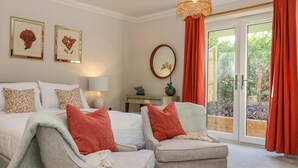 Un dormitorio con una cama, dos sillones, una mesita de noche, una lámpara, un espejo y una ventana con cortinas rojas.