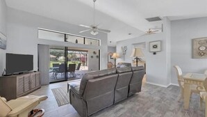 A spacious living room with a grey sofa, a television on a cabinet, and a dining area with a table and chairs.