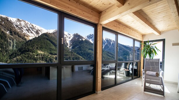 A modern interior with large windows offering mountain views, wooden ceiling beams, and a visible staircase. - Das Eppensteiner (Navis)