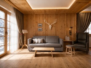 A cozy living room with a wooden ceiling, a grey sofa, a wooden coffee table, a leather armchair, and a deer skull mounted on the wall.