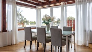 Un coin repas avec une table en verre et six chaises, de grandes fenêtres ornées de rideaux blancs et un plafond en bois.