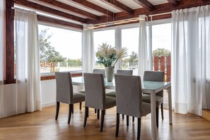 A dining area with a glass table and six chairs, large windows with white curtains, and a wooden ceiling.