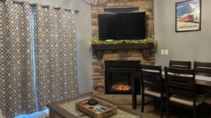 Uma sala de estar com lareira, uma televisão acima dela e uma área de refeições com cadeiras e uma mesa.