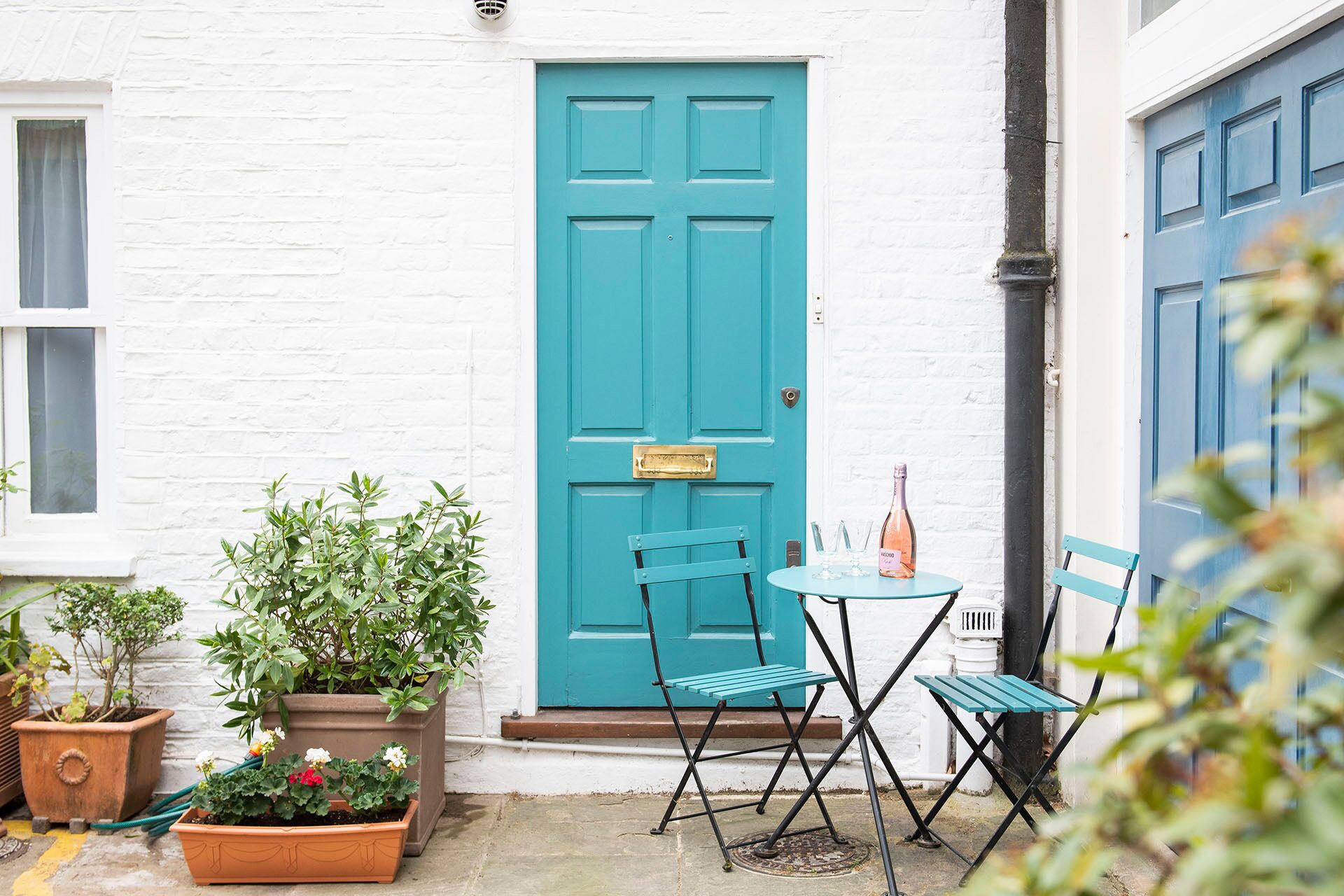 A white brick building with a turquoise door, a small table with chairs, and potted plants.