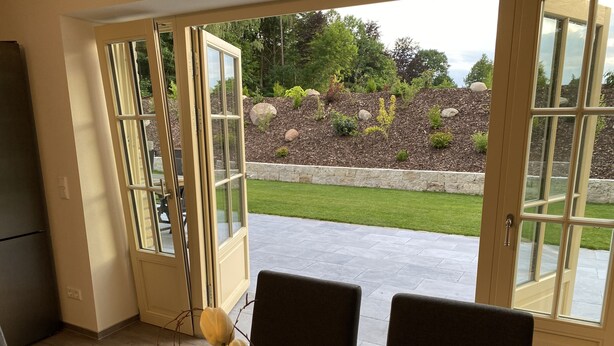 A dining area with a table and chairs, a vase with flowers, and a view of a landscaped garden through open French doors.