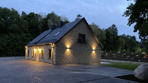 A stone building with a sloped roof and multiple windows, surrounded by a paved area and trees.
