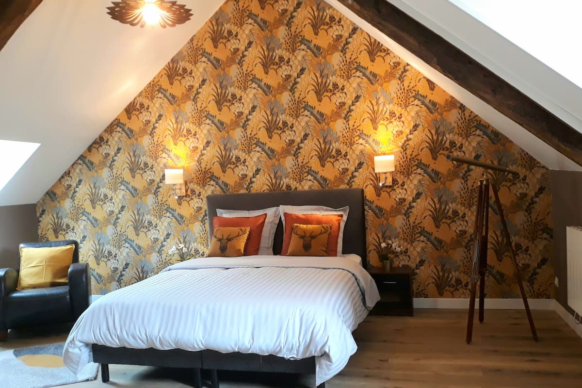 Une chambre avec un plafond en pente, un lit avec une literie blanche, deux lampes et une chaise avec un coussin jaune.