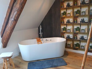 A modern bathroom with a freestanding bathtub, wooden shelves with plants, and a small stool.