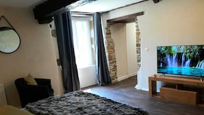 A bedroom with a bed, a TV on a wooden stand, a window with curtains, and a stone wall. - Le Manoir des Haies (Les Champs-Geraux)
