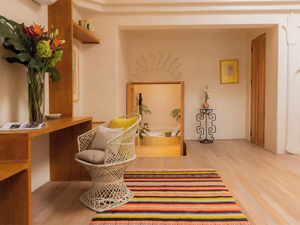 A room with a wicker chair, a colorful rug, a wooden desk, and a plant.