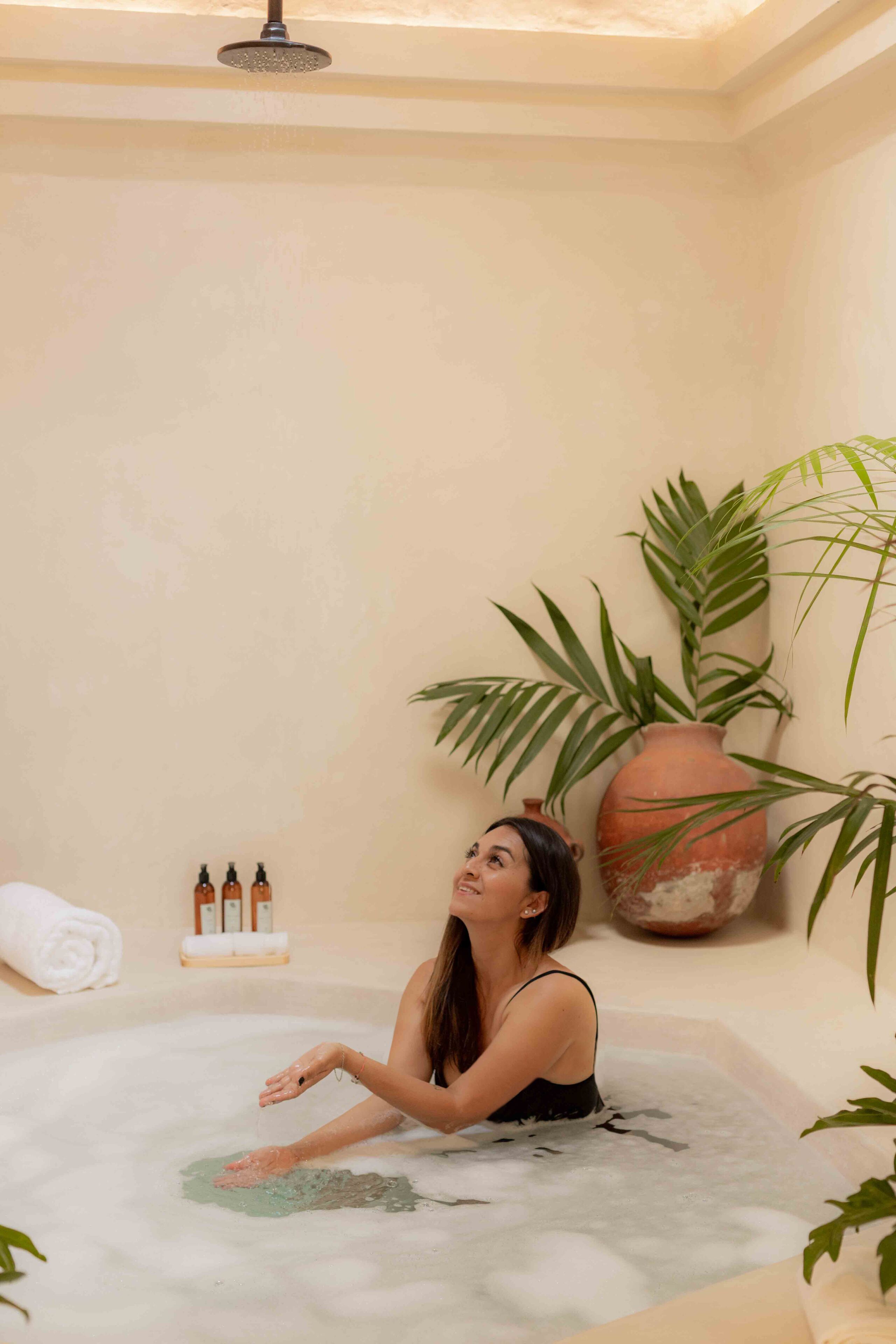 A person in a bathtub with a large potted plant and toiletries on the side.