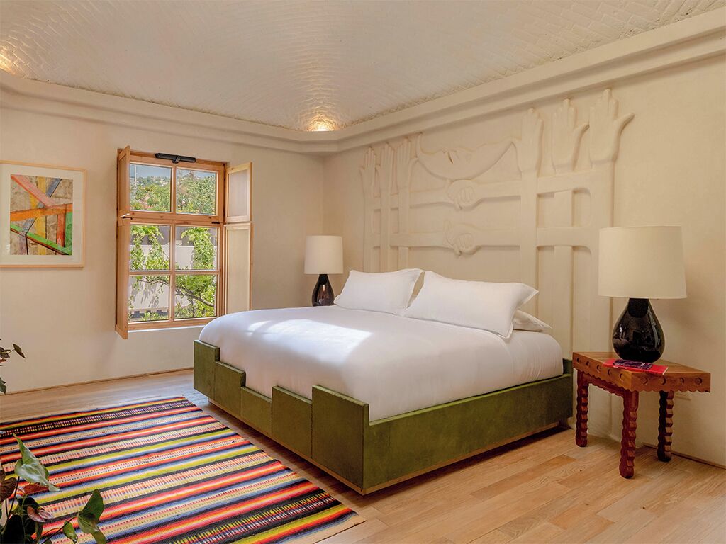A bedroom with a bed, two bedside lamps, a colorful striped rug, a window with a view of greenery, and a wall with a decorative panel.