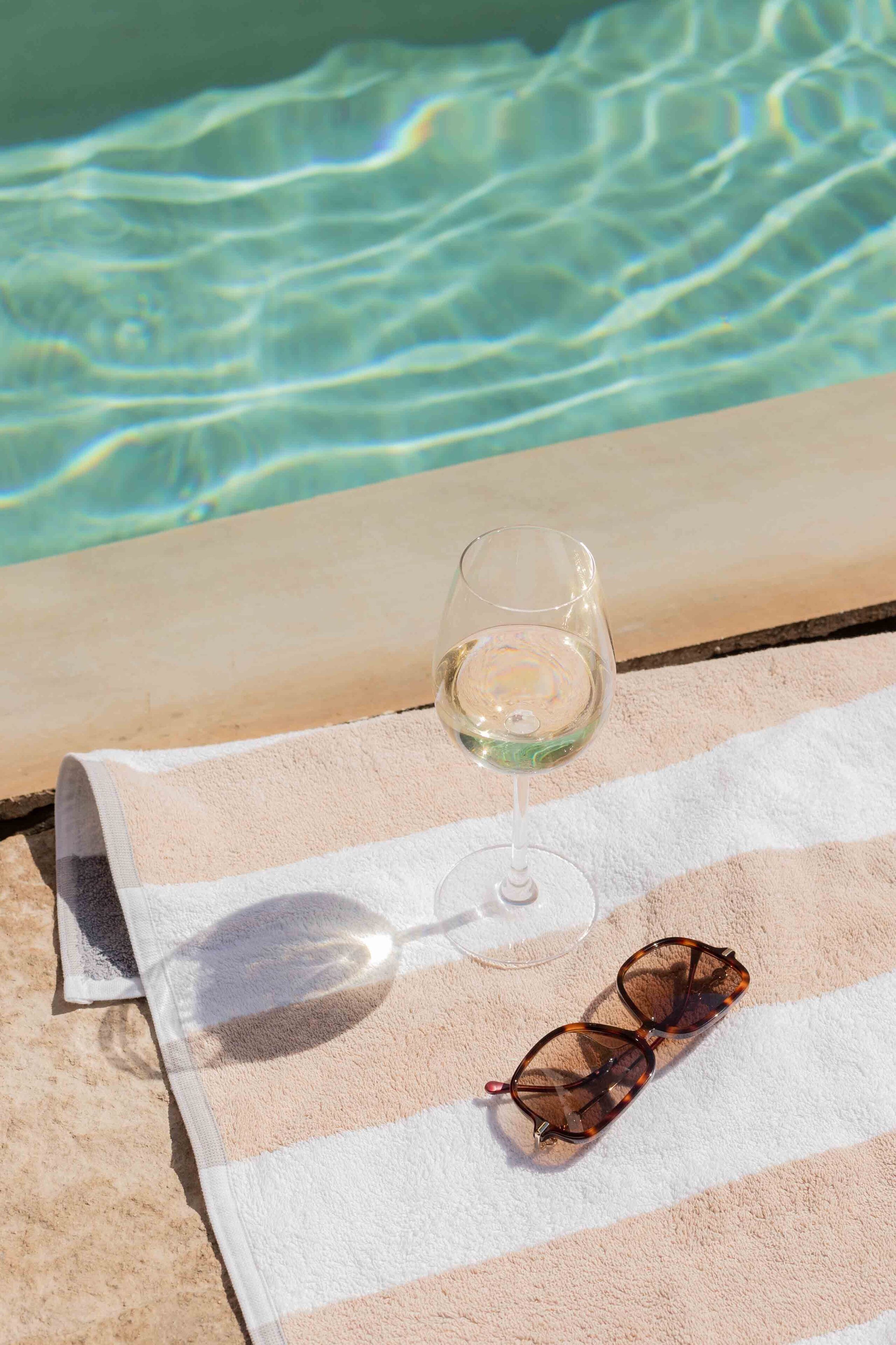 Una copa de vino blanco y gafas de sol sobre una toalla a rayas junto a una piscina.